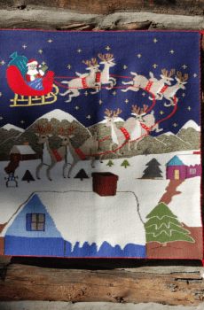 A decorative Christmas-themed tapestry featuring Santa Claus in a sleigh pulled by reindeer, flying over snowy houses, displayed on a wooden wall.