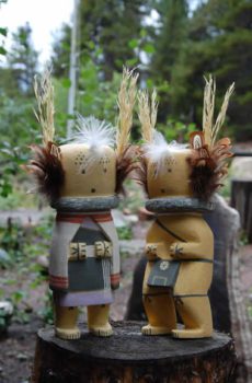 Two Yellow Cricket Mana (female, left) figurines with wheat heads, feather and fur details, standing on tree stumps in a woodland setting.