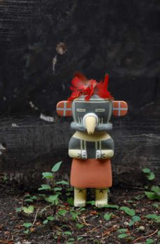 Colorful figurine of a Red Skirt Kachina character with human attributes, standing against a dark background, adorned with red feathers on its head.