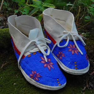 A pair of traditional American Indian Art beaded moccasins on a mossy surface, featuring vibrant blue fabric with red and yellow floral patterns.
