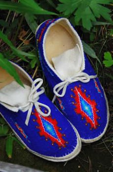 A pair of traditional blue moccasins with red and white bead patterns, placed on a grassy surface next to a wooden edge.