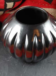 A polished metallic vase with a fluted design sits on a grey slate against a red patterned fabric background.