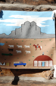 Sentence with Product Name: A colorful woven Revival depicting a rural scene with mountains, animals, a vehicle, and a building, hung on a wooden log wall.