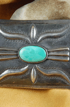 Ingot silver buckle with 9 conchos with a central turquoise stone and embossed floral designs, displayed on a beige fabric surface.